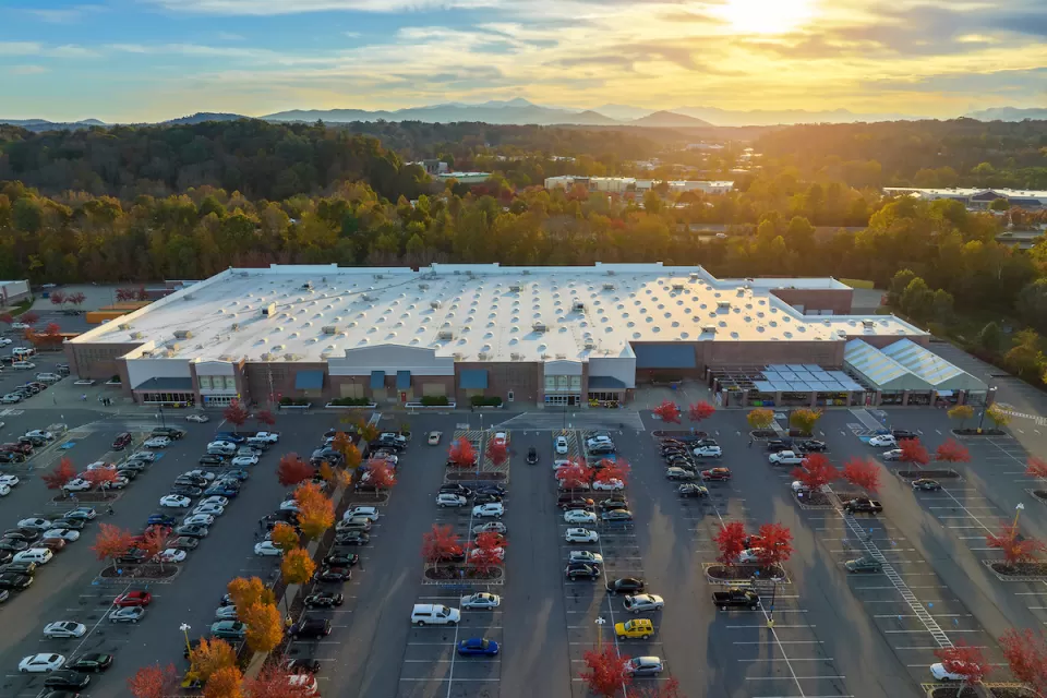 Aerial view of large parking lot in front of large grocery or big-box store with small fall-colored trees among parked cars and sunset sky in background.